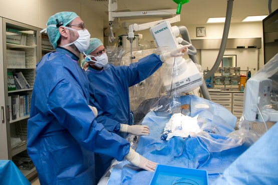 Zwei Ärzte in blauer OP-Kleidung und Mundschutz führen Eingriff am Bildgebungsgerät im sterilen Operationssaal durch