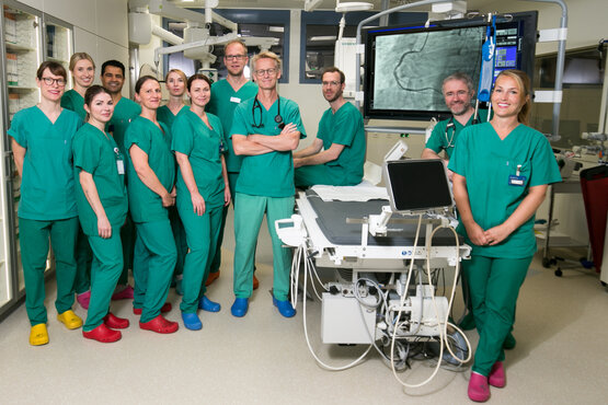 Zwölfköpfiges Team aus Ärzt:innen und Pflegepersonal in grüner OP-Kleidung und bunten Clogs im Herzkatheterlabor