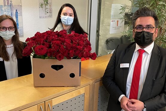 Zwei Frauen und ein Mann mit Masken am Empfang, großer Karton mit roten Rosen auf der Theke zum Weltfrauentag