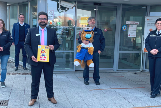 Fünf Personen vor Glastür, Mann hält „HILFE INSEL“-Schild mit Comic-Löwen, Polizist mit Plüschlöwen-Maskottchen