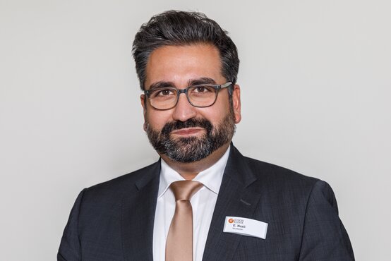 Person mit dunklem Anzug, weißem Hemd, beigefarbener Krawatte, Brille und Namensschild vor hellgrauem Hintergrund