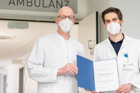 Zwei Ärzte, einer hält Urkunde zum Nachwuchspreis in Klinikflur