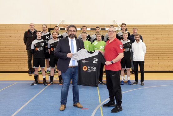 Gruppenfoto einer Handballmannschaft in Sporthalle; vorn Trikotübergabe mit Schön-Klinik-Logo, im Hintergrund Spieler vor Handballtor