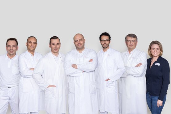 Siebenköpfiges Team der Chirurgie, sechs Männer in weißen Kitteln, eine Frau im Blazer, Gruppenfoto vor hellem Hintergrund