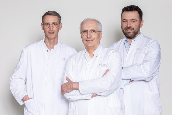 Drei Ärzte im weißen Kittel mit Namensschild stehen nebeneinander vor hellem Hintergrund und lächeln in die Kamera