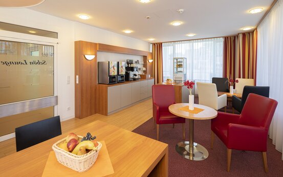 Helle Lounge der Schön Klinik Lorsch mit modernen Sitzmöbeln, Holztischen, bodentiefen Fenstern, Selbstbedienungs-Theke und frischem Obst
