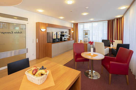 Helle Lounge der Schön Klinik Lorsch mit modernen Sitzmöbeln, Holztischen, bodentiefen Fenstern, Selbstbedienungs-Theke und frischem Obst