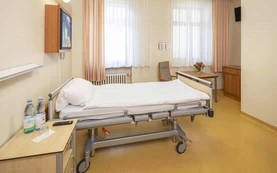 Helles Einzelzimmer der Schön Klinik Lorsch mit Pflegebett, Nachttisch, Tisch, Schrank, Fernseher und großem Fenster