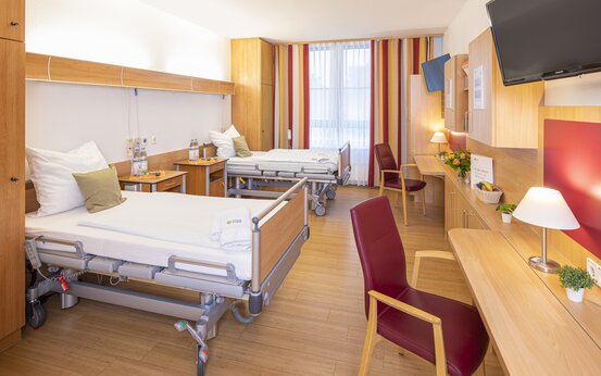 Modernes Komfortzimmer mit zwei Pflegebetten, Holzfußboden, Schreibtisch, roten Stühlen, Blumen, Obst und großem Fenster