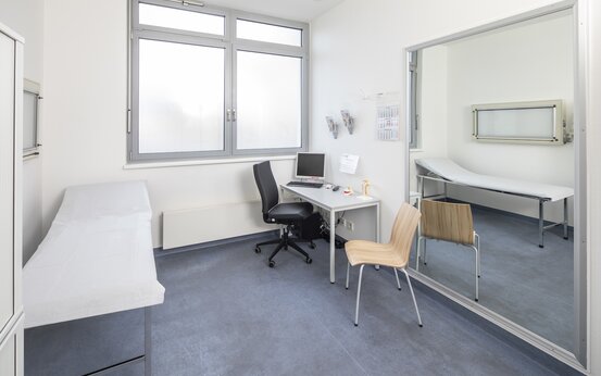 Heller Untersuchungsraum mit Liege, Schreibtisch, Computer, großem Fenster und blauem Boden in der Schön Klinik Lorsch
