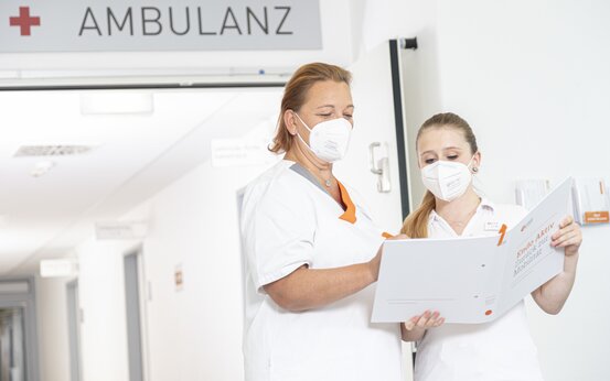 Zwei Pflegekräfte mit FFP2-Masken besprechen Patientenakte im Flur der Ambulanz, Teamarbeit in moderner Klinik