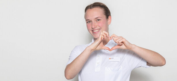 Lächelnde Pflegekraft in weißer Dienstkleidung formt mit beiden Händen ein Herz vor der Brust