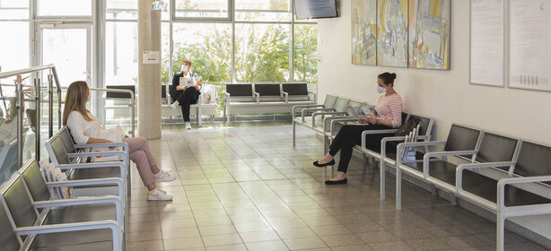 Heller Wartebereich mit grauen Metallbänken, drei Personen mit Masken lesen Zeitschriften, große Fenster, Wandbilder