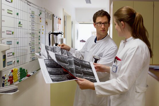 Zwei medizinische Fachkräfte in weißen Kitteln betrachten gemeinsam mehrere Röntgenbilder vor einer Wandtafel mit Magneten und Notizen