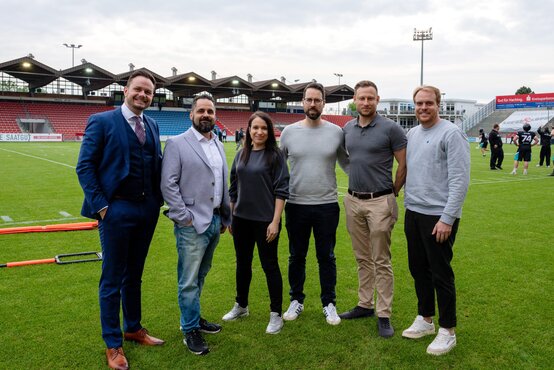 Sechs Personen stehen nebeneinander auf dem Footballfeld im Stadion, rote und blaue Sitzreihen, weitere Menschen trainieren im Hintergrund