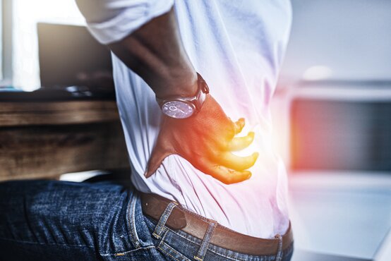 Person sitzt auf Stuhl, trägt Jeans und weißes Hemd, fasst sich mit rechter Hand an den schmerzenden unteren Rücken