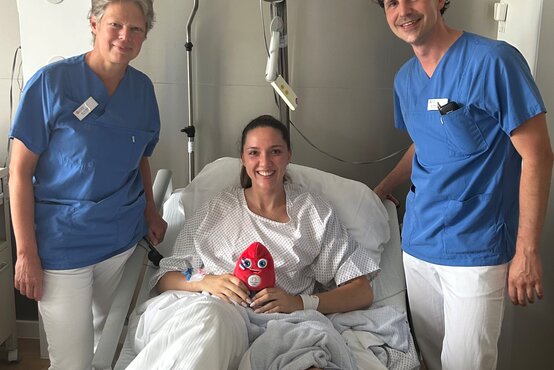 Patientin mit Gipsbein im Krankenbett, hält rotes Stofftier, betreut von zwei Klinikmitarbeitenden