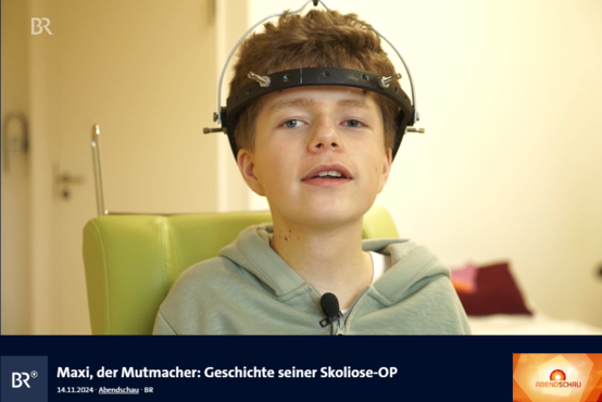 Junger Mensch mit Fixateur externe am Kopf sitzt auf grünem Stuhl, lächelt in hellem Raum nach Skoliose-OP