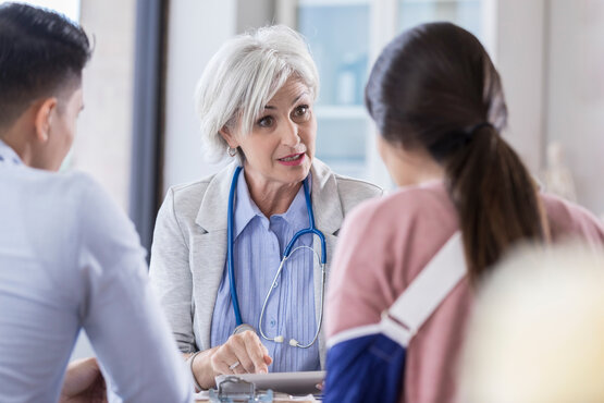 Ärztin mit Stethoskop spricht mit zwei jungen Erwachsenen, eine Person mit blauer Armschlinge im Untersuchungszimmer