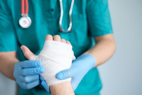 Arzt in grüner OP-Kleidung legt mit blauen Handschuhen einen weißen Verband an eine verletzte Patientenhand an