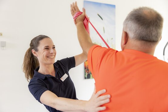Physiotherapeutin unterstützt männlichen Patienten bei Schulterübung mit rotem Trainingsband
