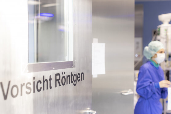 Edelstahl-Tür mit Sichtfenster und Aufschrift „Vorsicht Röntgen“, dahinter Person in blauer OP-Kleidung