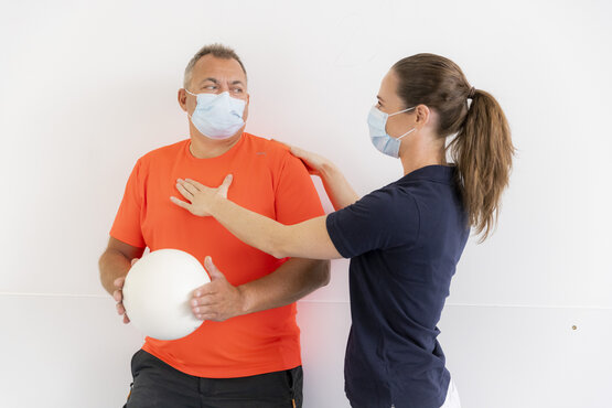 Physiotherapeutin und Maske hilft einem Patienten mit Ball und Maske bei Therapeutischen Übungen