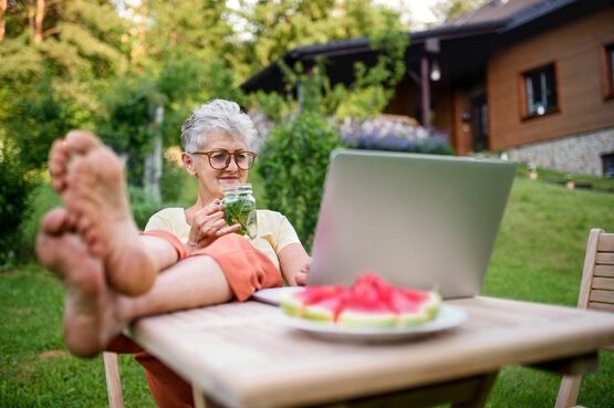 Ältere Frau mit grauen Haaren sitzt barfuß entspannt im Garten am Holztisch mit Laptop und Getränk