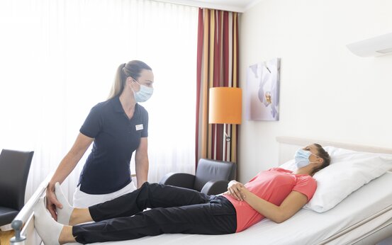 Pflegekraft in blauem Shirt hält die Beine einer Patientin im Bett, beide tragen Masken, heller Raum mit Fenster