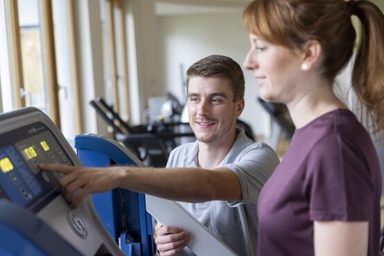 Junger Therapeut erklärt einer Frau im Fitnessstudio die Bedienung eines modernen Fitnessgeräts und zeigt auf das Display