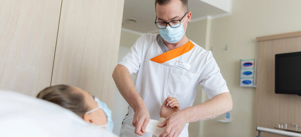 Krankenpfleger mit Brille und Maske legt Patientin im Krankenhauszimmer einen Verband am Arm an, beide tragen medizinische Masken