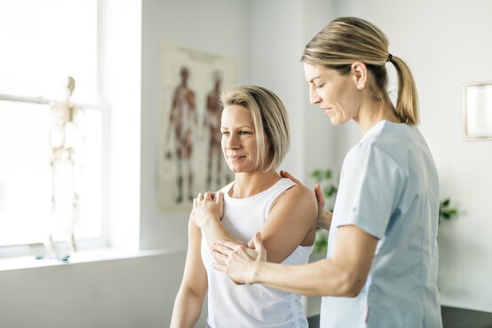 Physiotherapeutin untersucht und behandelt Schulter einer Patientin in heller Praxis, anatomisches Poster und Skelettmodell im Hintergrund
