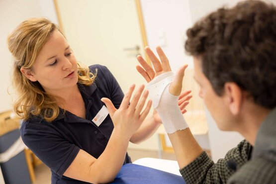 Therapeutin im dunkelblauen Poloshirt erklärt Handübung, Patientin mit weißer Handschiene am Tisch im hellen Therapieraum
