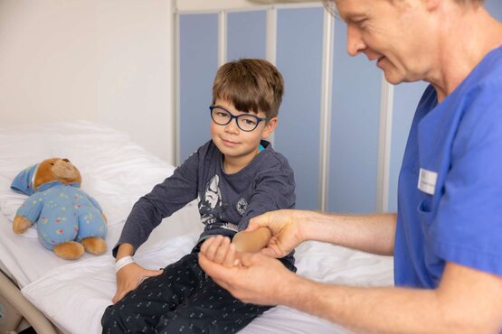 Physiotherapeut behandelt den Arm von einem Kind mit Brille auf Krankenhausbett in heller Kinderstation