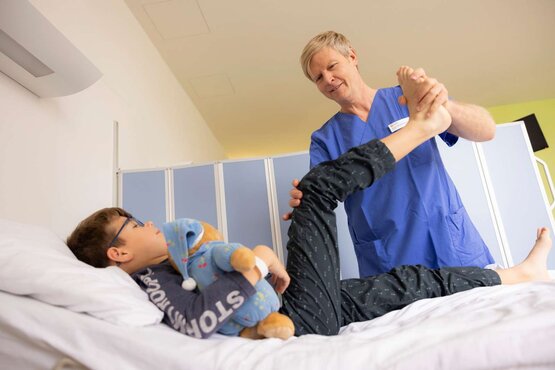 Physiotherapeut behandelt Kind mit Brille und Kuscheltier auf Krankenhausbett in heller Kinderstation