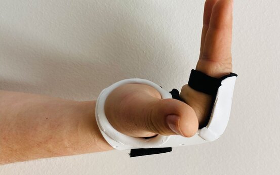 Hand mit weißer Handorthese vor heller Wand, Mittel- und Zeigefinger aufrecht, Daumen abgespreizt