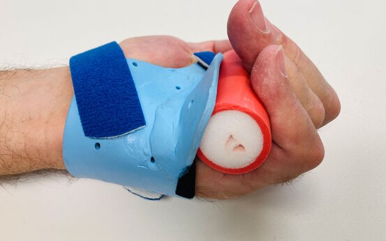 Nahaufnahme einer Hand mit blauer Handgelenkschiene und Klettverschluss, die einen rot-weißen Schaumstoffgriff hält