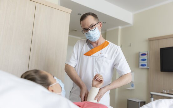 Pfleger mit Mundschutz untersucht Arm einer Patientin mit Gips im Krankenhauszimmer