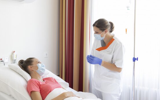 Medizinische Fachkraft mit Schutzmaske und Handschuhen betreut Patientin im rosa T-Shirt im Krankenhausbett mit Infusion