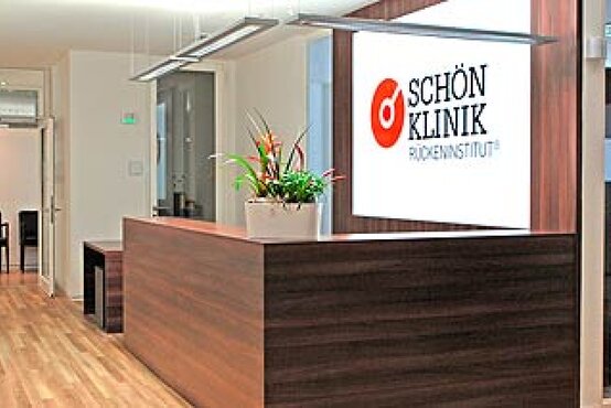 Heller Empfangsbereich der Schön Klinik Rückeninstitut mit Holzfußboden, moderner dunkler Empfangstheke, Blumenkübel und beleuchtetem Logo