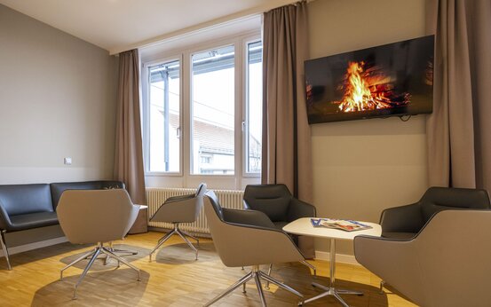 Heller Aufenthaltsraum mit großen Fenstern, grauen Sesseln um weiße Tische, schwarzes Sofa, Fernseher mit Kaminfeuer