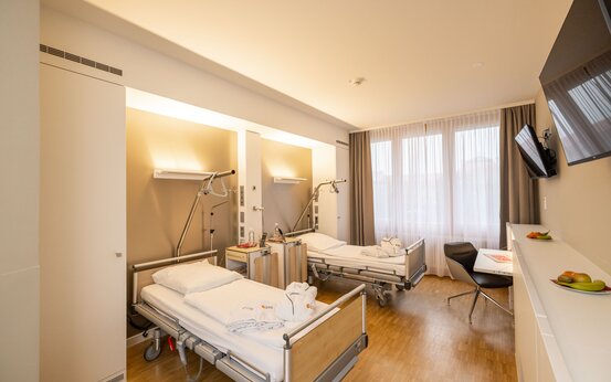 Modernes, helles Zweibett-Patientenzimmer mit verstellbaren Betten, Schreibtisch, Flachbildfernseher und Obstschalen