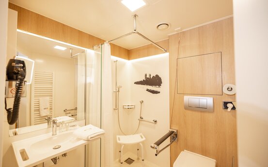 Barrierefreies Badezimmer mit begehbarer Dusche, Duschhocker, Haltegriffen, Toilette und großem Spiegel