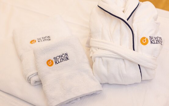 Zwei gefaltete weiße Handtücher und ein Bademantel mit Schön Klinik Logo auf weißem Bettlaken