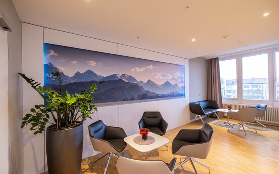 Moderner, heller Aufenthaltsraum mit großen Fenstern, Sitzgruppen aus schwarzen und grauen Sesseln, Panoramabild und Zimmerpflanze