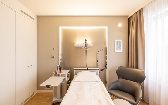 Einzelzimmer in der Klinik mit komfortablem Krankenbett, Nachttisch, Sessel und großem Fenster