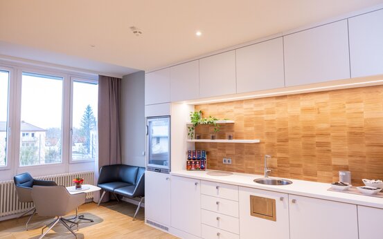 Patienten Küche mit weißer Einbauküche, Holzrückwand, Edelstahlspüle, zwei Sesseln und Beistelltisch mit Blumenstrauß