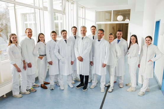 Zwölf Personen in weißen Kitteln und medizinischer Kleidung stehen lächelnd im hellen Klinikflur mit großen Fenstern