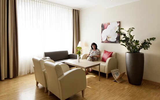 Frau liest Zeitschrift auf hellem Sofa in modernem Wartebereich mit Sesseln, Holztisch, Vase mit Blumen und großer Pflanze