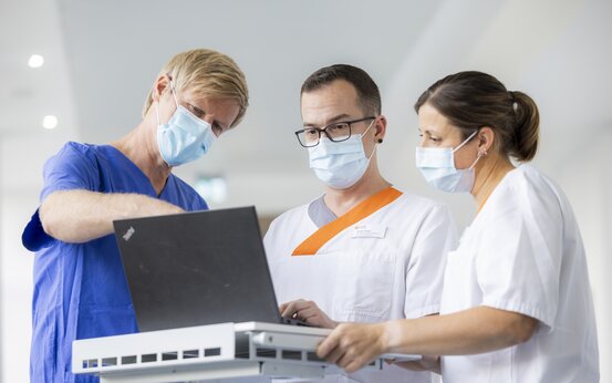 Drei Personen in medizinischer Kleidung und Masken stehen an einem Laptop auf Rollwagen, eine Person zeigt auf den Bildschirm